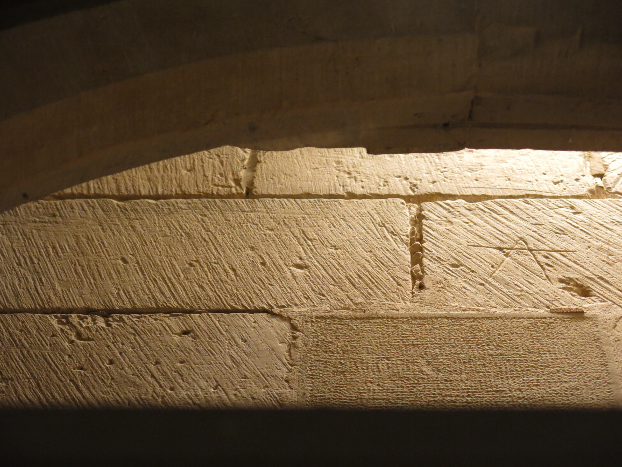 Parallel chisel marks and a stonemason mark shaped as a crossed-out inverted V, possibly an A or a compass, on the wall of the church.