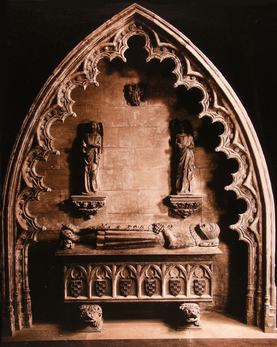 Black and white archival photograph of the tomb in full armor in an antique dealers' atelier. The effigy is displayed under a free-standing stone arch with scalloped edges and small carved shields along its borders.