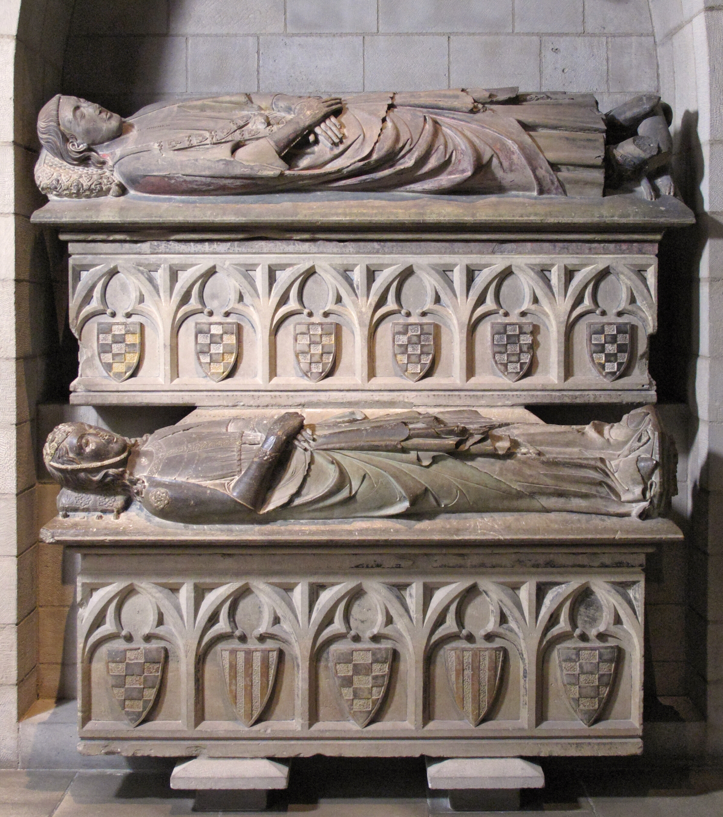 Two stacked tomb effigies of a female and male figure, husband and wife, each lying over a sarcophagus with coat of arms. The ensemble is carved in limestone with polychromy.
