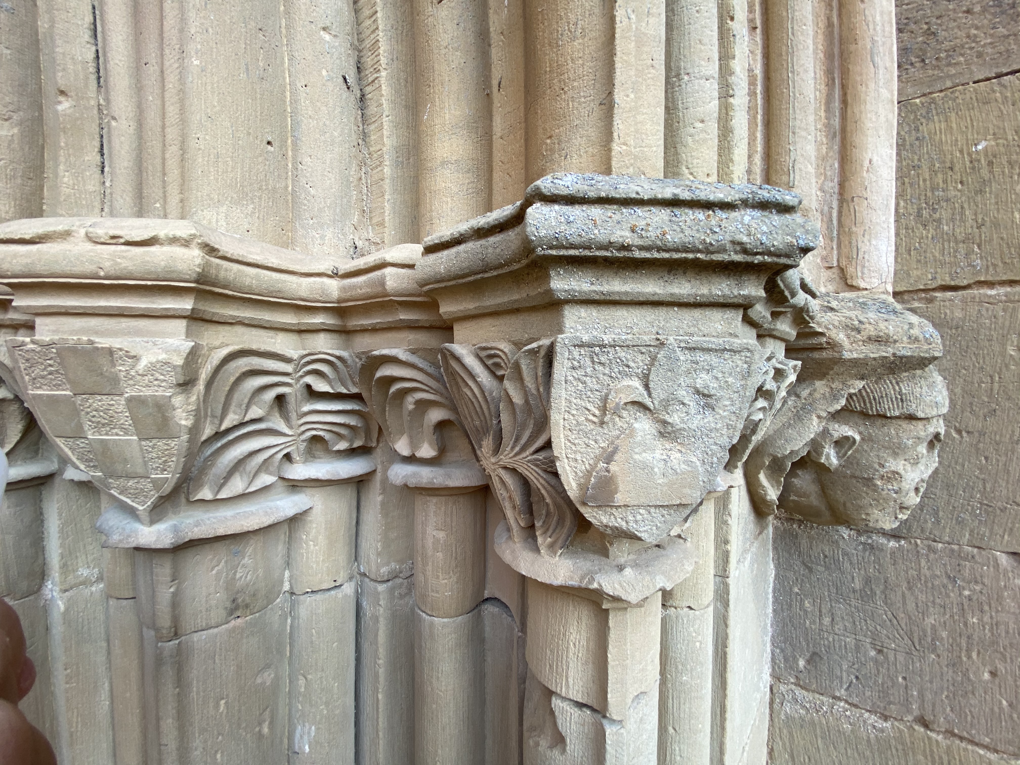 Series of stone capitals with coat of arms, foliate motifs, a flowering hill, and a small head bracketing the gothic doorway on the right.