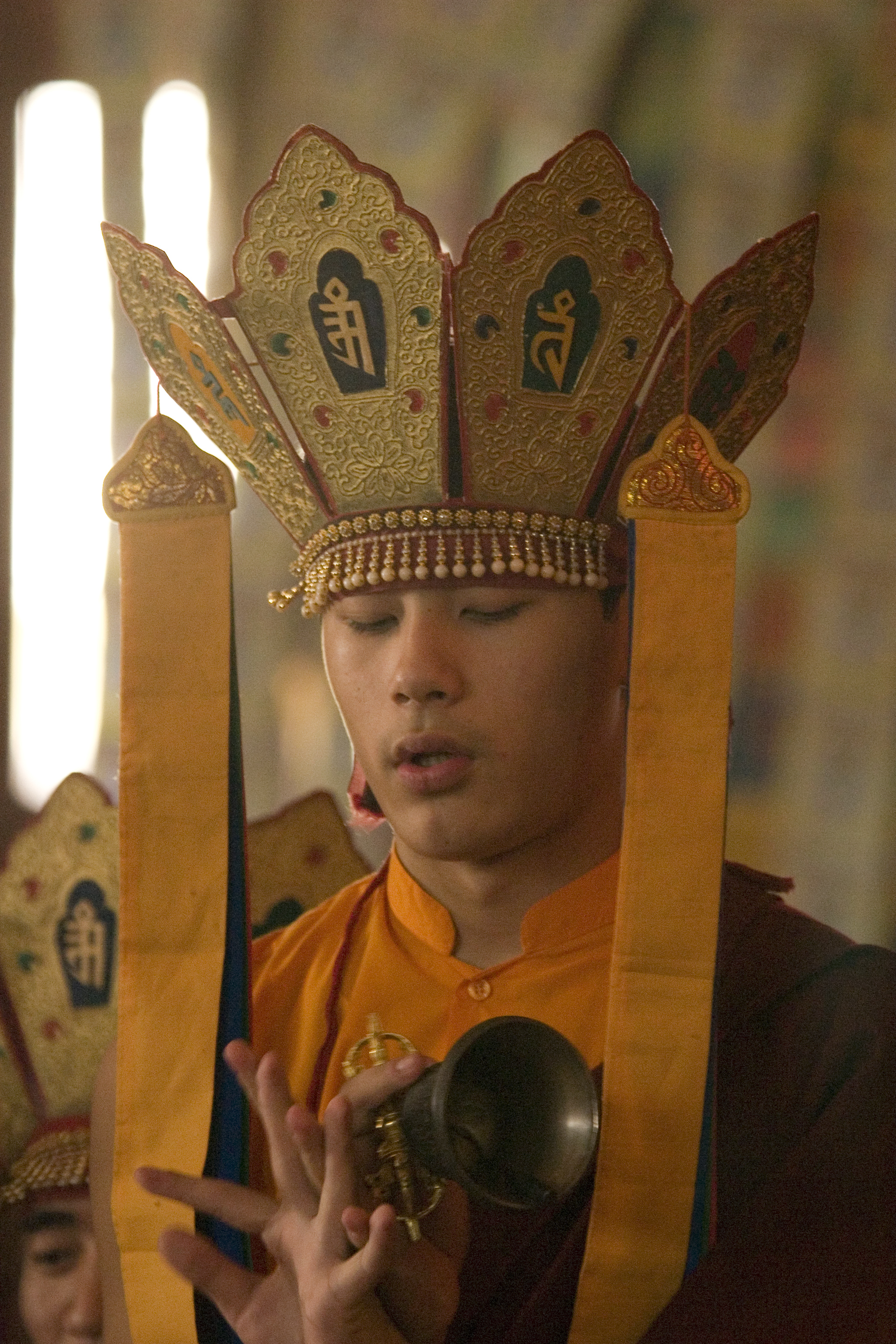 Young Tibetan lama wearing a robe and five paneled textile crown that splays outward with yellow ribbons hanging down either side.